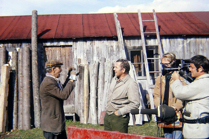 Photo de Grand-Louis Harvey discutant avec Pierre Perrault devant une grange. Derrière Perrault se tient un caméraman et un preneur de son.