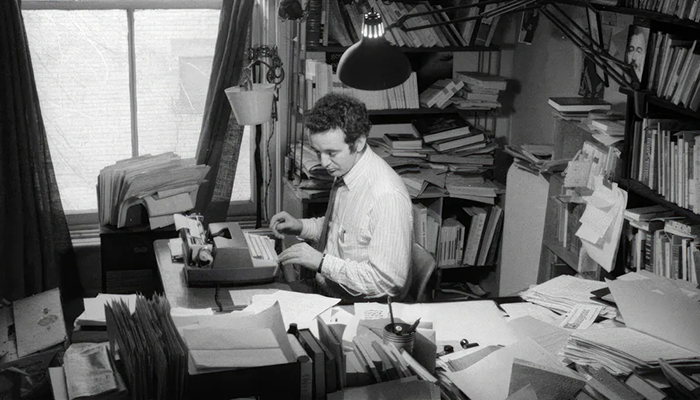 Photo de Gérald Godin assis à son bureau devant sa machine à écrire dans une pièce très encombrée de papiers et différentes choses.