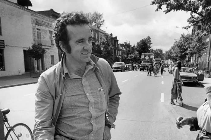 Photo de Gérald Godin, souriant, dans une rue libérée pour laisser passer un défilé.