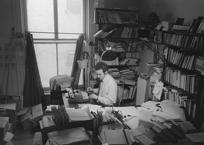 Photo de Gérald Godin tapant à la machine dans une pièce très encombrées de paperasse.
