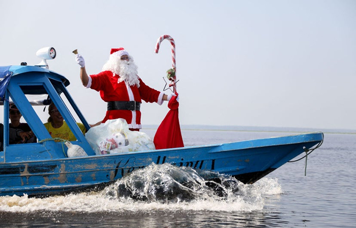 Photo en couleurs. Un homme habillé en père Noël se tient sur le devant d'un petit esquif naviguant sur la mer.