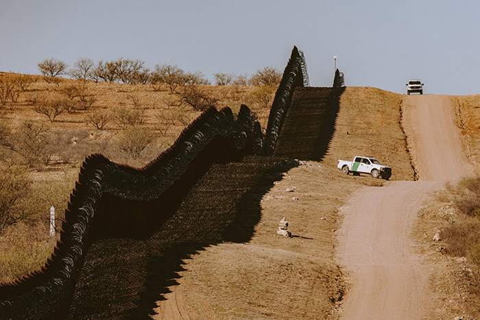 Photo d'un long mur métallique dans le désert de l'Arizona, longé d'une route de terre où est stationné un camion pick-up.