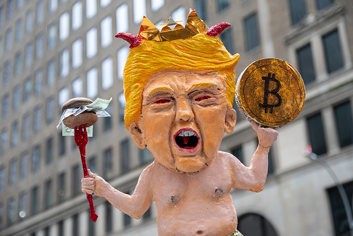 Photo d'une effigie caricaturant Trump le montrant nu, portant une couronne, une fourchette tenant un hamburger plein d'argent en guise de sceptre dans une main et une pièce avec le symbole de Bitcoin dans l'autre.