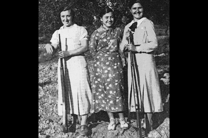 Vieille photo de trois femmes debout et souriantes. Deux d'entre elles tiennent un fusil.